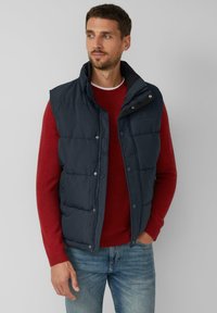 Granatowa kamizelka puchowa z wysokim kołnierzem, zapięciem na napy z przodu i bocznymi kieszeniami, noszona na czerwonym swetrze. Gładka tekstura, casualowy design.