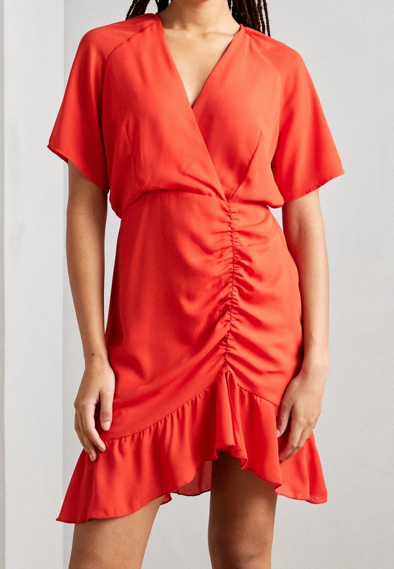 Femme portant une robe portefeuille rouge vif à manches courtes, devant froncé et ourlet asymétrique à volants, debout devant un fond uni.