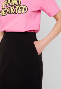 T-shirt di cotone rosa con testo "FAINT HEARTED", abbinata a una gonna nera a vita alta con tasche laterali e una texture liscia.