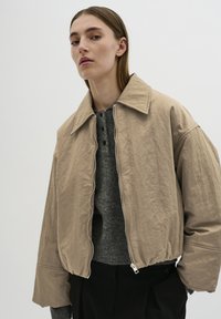 Beige kort jacka med stor krage, dragkedjestängning och lös passform på ärmarna, bärs över en grå stickad topp med knappar.