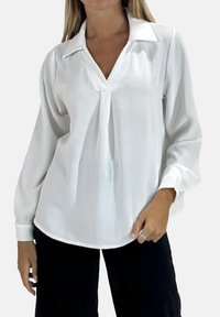 Blouse blanche à manches longues avec un col chemise, tissu texturé et coupe décontractée. Présente un léger pli à l'avant.