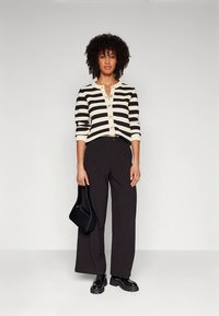 Zwart-wit gestreepte cardigan met ronde gouden knopen, gecombineerd met hooggetailleerde zwarte broek. Zwarte handtas en veter schoenen maken de look compleet.