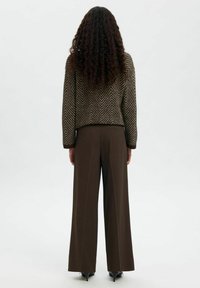 Personne aux longs cheveux bouclés portant un pull marron à motifs, un pantalon marron à jambes larges et des chaussures à talons noires, vue de dos sur un fond blanc uni.