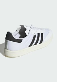 Białe sneakersy z czarnymi paskami, teksturowana cholewka z płótna, gumowa podeszwa i wyściółka w okolicy kostki. Logo "VELOSAMBA" na pięcie.
