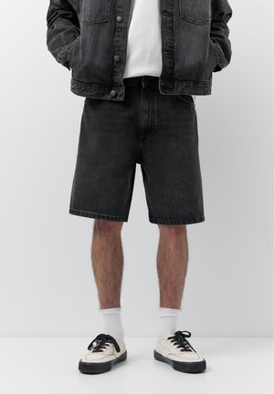 Person iført sorte cowboyshorts, sort cowboyjakke, hvide sokker og hvide sneakers med sorte snørebånd stående mod en hvid baggrund.