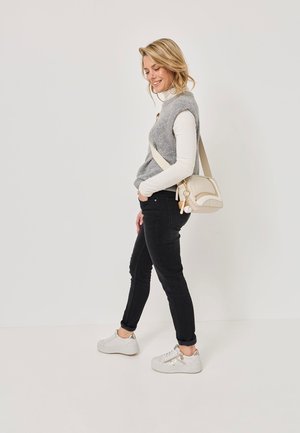 Mujer sonriendo, caminando con un bolso bandolera beige, vistiendo un suéter sin mangas gris, una camisa de manga larga blanca, jeans negros y zapatillas blancas.