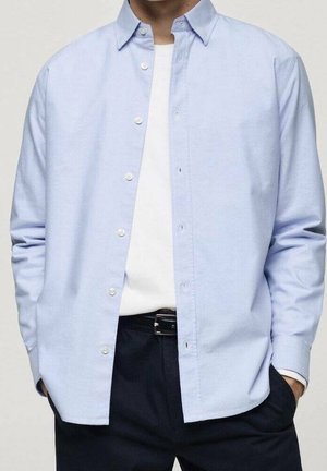 Homme portant une chemise bleu clair boutonnée par-dessus un t-shirt blanc, avec un pantalon foncé et les mains dans les poches, torse visible.