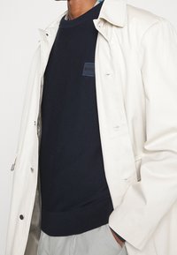 Granatowy sweter o teksturowanej powierzchni z czarną naszywką z logo pod kremowym, lekkim płaszczem. Widoczne szare spodnie wykonane z gładkiego materiału.