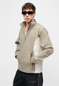 Νεαρός άνδρας που φοράει μπεζ μπουφάν με μισό φερμουάρ και πλαϊνές τσέπες και μαύρο παντελόνι New Balance, ποζάροντας μπροστά σε απλό ανοιχτό φόντο.