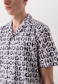 Lila anyagú, rövid ujjú ing, amelyen merész fekete grafikai mintázat látható, ismétlődő "HUGO" felirattal különböző méretekben. Gombos gallér kialakítás.