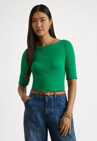 Πράσινη μπλούζα με τρία τέταρτα μανίκι, λεία υφή, λαιμόκοψη καραβάκι, συνδυασμένη με τζιν ψηλόμεσο τζιν και καφέ ζώνη με χρυσές λεπτομέρειες.
