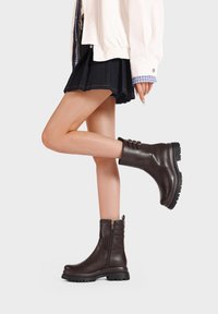 Stivaletti alla caviglia in pelle marrone con texture liscia, cerniera laterale e tre cinturini decorativi sul gambale. Suola spessa in gomma per una migliore aderenza.