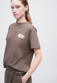 Sieviete, ģērbta brūnā īsām piedurknēm krekliņā un atbilstošās biksēs ar baltu "ellesse" logo, stāv ar vienu roku kabatā uz vienkrāsainas fona.