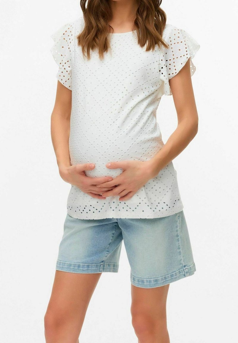 Femme enceinte tenant son ventre, portant un chemisier blanc à broderies anglaises avec des manches volantées et un short en jean bleu clair.