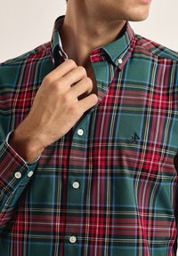 Camisa a cuadros en verde y rojo con cuello abotonado, botones blancos y detalle de logotipo, que presenta una textura suave de algodón y rayas en contraste.