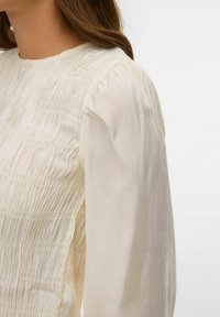 Krēmkrāsas blūze ar teksturētu, plīsētu augšdaļu, apaļu izgriezumu un garām, gludām piedurknēm, izceļot vieglu audumu un smalku šuvumu.
