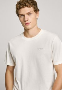 Light beige cotton t-shirt with short sleeves, crew neck, and a subtle logo printed on the chest in a darker shade. Smooth texture.