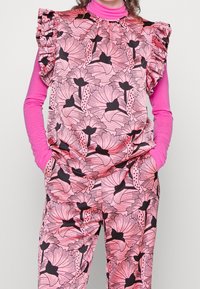 Růžový outfit s květinovým vzorem, který obsahuje jasně zbarvený top s dlouhým rukávem pod krátkým rukávem s volány na ramenou.