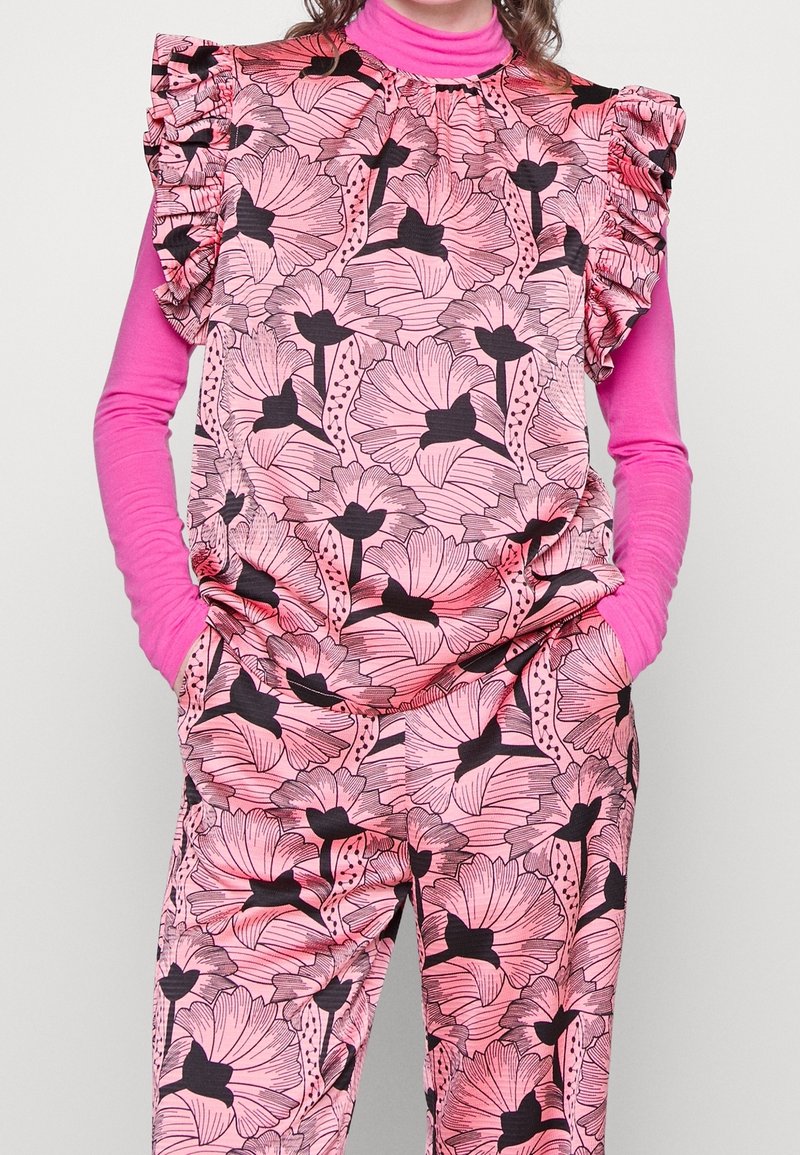 Růžový outfit s květinovým vzorem, který obsahuje jasně zbarvený top s dlouhým rukávem pod krátkým rukávem s volány na ramenou.