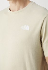 T-shirt en coton beige avec un col rond, présentant un petit logo blanc sur la partie gauche de la poitrine. Texture douce, manches courtes, design simple.