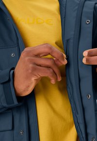 Veste matelassée navy avec boutons-pression, zips et poches latérales, superposée sur une chemise jaune avec logo de marque en relief. Mains ajustant la veste.