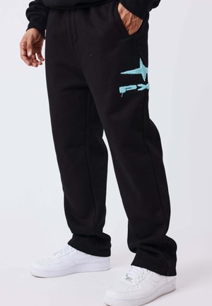 Personne portant un pantalon de survêtement noir avec un motif bleu clair sur la cuisse et des baskets blanches, debout avec une main dans la poche.
