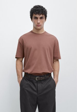 Giovane uomo con capelli ricci che indossa una maglietta marrone a maniche corte infilata in pantaloni grigi con cintura nera, in piedi con le mani nelle tasche.