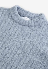 Ljust grå stickad tröja med en strukturerad, ribbad mönstring, rundad hals, tillverkad av mjukt, fluffigt material. Varumärkesetikett synlig.