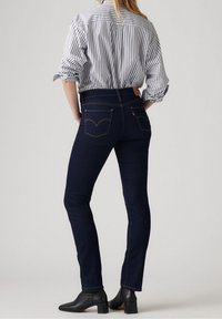 Jeans de mezclilla azul oscuro con una forma ajustada, que presentan bolsillos traseros con un diseño curvado y costuras naranjas. Combinados con una camisa a rayas.