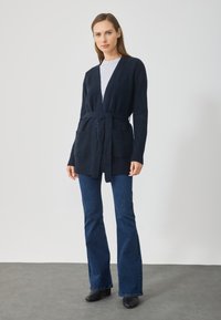 Námornícky pletený kardigán s opaskom, v tvare V, rebrované manžety a dve predné vrecká, nosený cez svetlo šedý top a rozšírené tmavé džínsy.