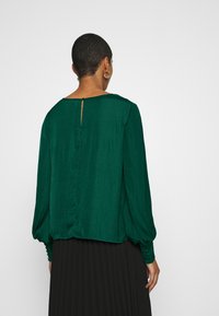 Blusa verde a maniche lunghe realizzata in tessuto morbido. Caratterizzata da una scollatura a forma di chiave sul retro, vestibilità ampia, maniche a sbuffo e dettagli con bottoni sui polsini.