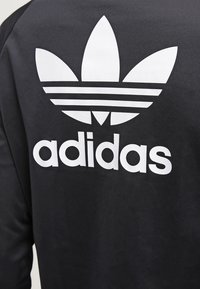Felpa nera in tessuto con il grande logo bianco di Adidas, con design a trifoglio e tre strisce orizzontali sopra il nome del marchio.