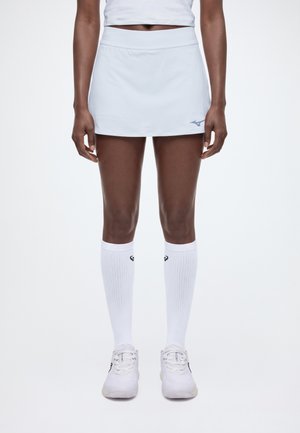 Un mannequin se tient devant un fond blanc, vêtu d’une jupe de sport blanche, d’un haut court, de chaussettes montant jusqu’aux genoux et de chaussures de tennis.