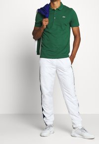 Grön pikétröja med krage, vita träningsbyxor med marinblå sidostripes och ljusgrå sportskor, med en lila tröja hängande över armen.