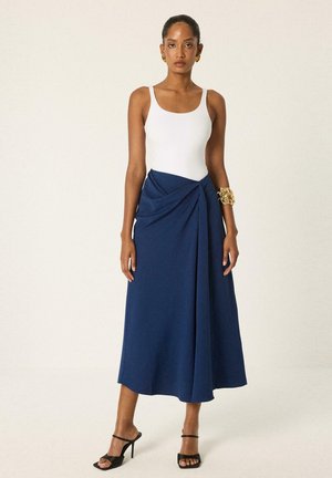 Donna che indossa un top bianco senza maniche, gonna midi blu navy drappeggiata, sandali neri con tacco alto, bracciale dorato e orecchini dorati su sfondo uniforme.