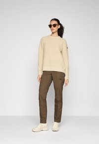 Beige gebreide trui, bruine cargobroek met zijzakken en lichte sneakers. Model draagt een paar bruine zonnebril; haar in een losse paardenstaart.