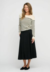 Langer, schwarzer, plissierter Rock kombiniert mit einem cremefarbigen und schwarzen gestreiften Langarmshirt. Modell trägt schwarze Slipper und steht vor einem neutralen Hintergrund.