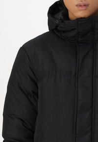 Zwarte pufferjack met een hoge kraag, voorzien van een gladde textuur en subtiele stikseldetails. Het heeft een rits aan de voorkant en verstevigde naden.