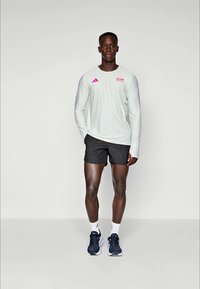 Männliche Sportbekleidung mit einem hellgrünen Langarmshirt mit rosa Akzenten und schwarzen Shorts, ergänzt durch marineblaue Turnschuhe.