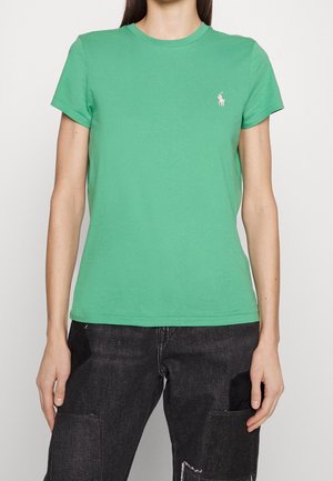 Femme portant un t-shirt vert uni à manches courtes avec un petit logo blanc sur la poitrine gauche, associé à un jean patchwork noir.