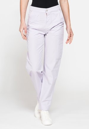Persona che indossa pantaloni dritti viola chiaro con tasca laterale e sneakers bianche, in piedi davanti a uno sfondo bianco uniforme.