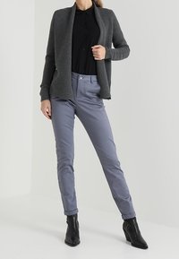 Grå stickad öppen kofta över en svart skjorta med knappar, kombinerad med ljusgrå byxor och svarta ankelstövlar. En enkel, figursydd design.