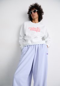 Sweatshirt branca com um logótipo vermelho da Nike, combinado com calças de fato largas em lavanda, adornadas com um pequeno logótipo roxo. O modelo usa óculos de sol brancos.