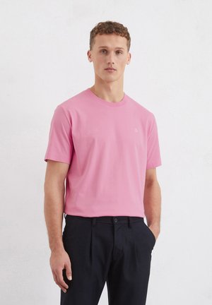 Pink cotton t-shirt with short sleeves and a round neckline, featuring a small embroidered logo on the chest. Paired with dark trousers.