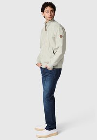 Mies, jolla on kiharat hiukset, pukeutuneena vaalean beigeen vetoketjulliseen takkiin, tummansinisiin farkkuihin ja valkoisiin lenkkareihin, seisoo kädet taskuissa.