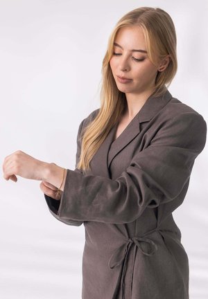 Braunes Leinengewand mit Taillengürtel und weiten Ärmeln, getragen von einer Person, die ein goldenes Armband am Handgelenk anlegt. Einfaches, strukturiertes Design.
