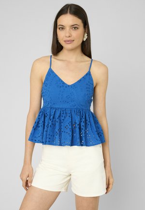 Femme portant un chemisier bleu sans manches à œillets avec un ourlet volanté et un short blanc, debout devant un fond gris uni.