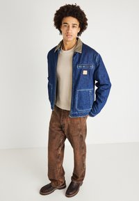 Jachetă din denim cu guler bej, fermoar frontal și două buzunare mari; asortată cu un pulover bej și pantaloni maro. Pantofi întunecați, texturizați, completează look-ul.