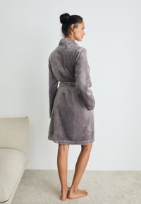 Kvinna som står barfota på en matta iförd en grå sammetrock med bälte, vänd åt sidan nära en beige soffa mot en enkel vit vägg.