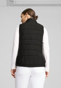 Svart pufferväst med horisontell sömmar, hög krage och sidofickor. Bärs över en vit långärmad skjorta som visar en slät textur.